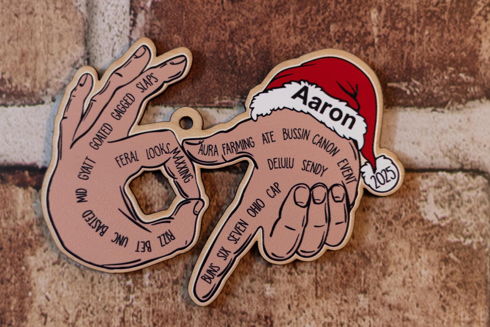 Personalized ‘67’ ornament with the name Aaron printed on the Santa hat. UV printed design shows illustrated hands forming 67 with modern slang words across the fingers. Brick wall backdrop.