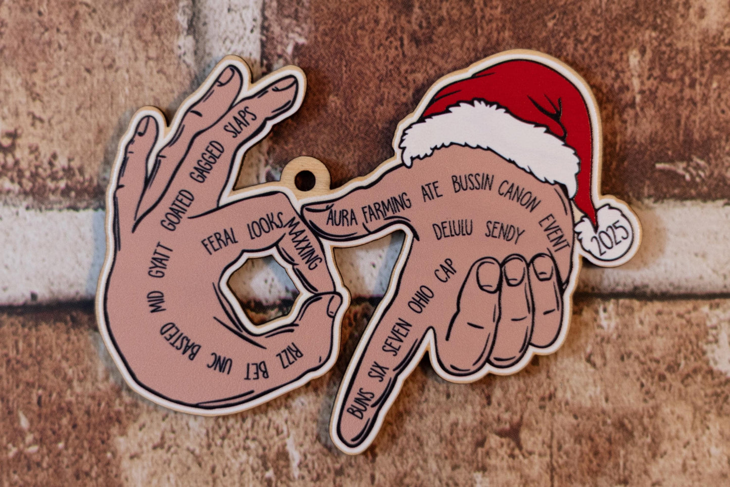 UV printed ‘67’ ornament featuring illustrated hands forming the number 67 with Gen Z slang words written across the fingers. Includes a Santa hat and small ‘2025’ tag. Displayed against a brick background.