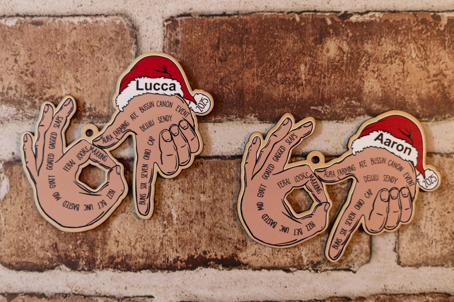 Two personalized ‘67’ ornaments shown side by side. One labeled Lucca and the other Aaron. Each ornament displays hands forming the number 67 with youth slang words printed on the fingers and a Santa hat with ‘2025.’ Set against a brick background.